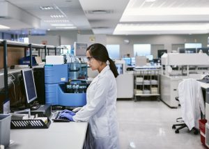 A focused female scientist working at a computer terminal in a modern, well-equipped laboratory. She is wearing protective eyewear and gloves, indicative of a safe working environment. The background is filled with advanced analytical instruments, suggesting a setting that prioritizes cutting-edge research and technological innovation.